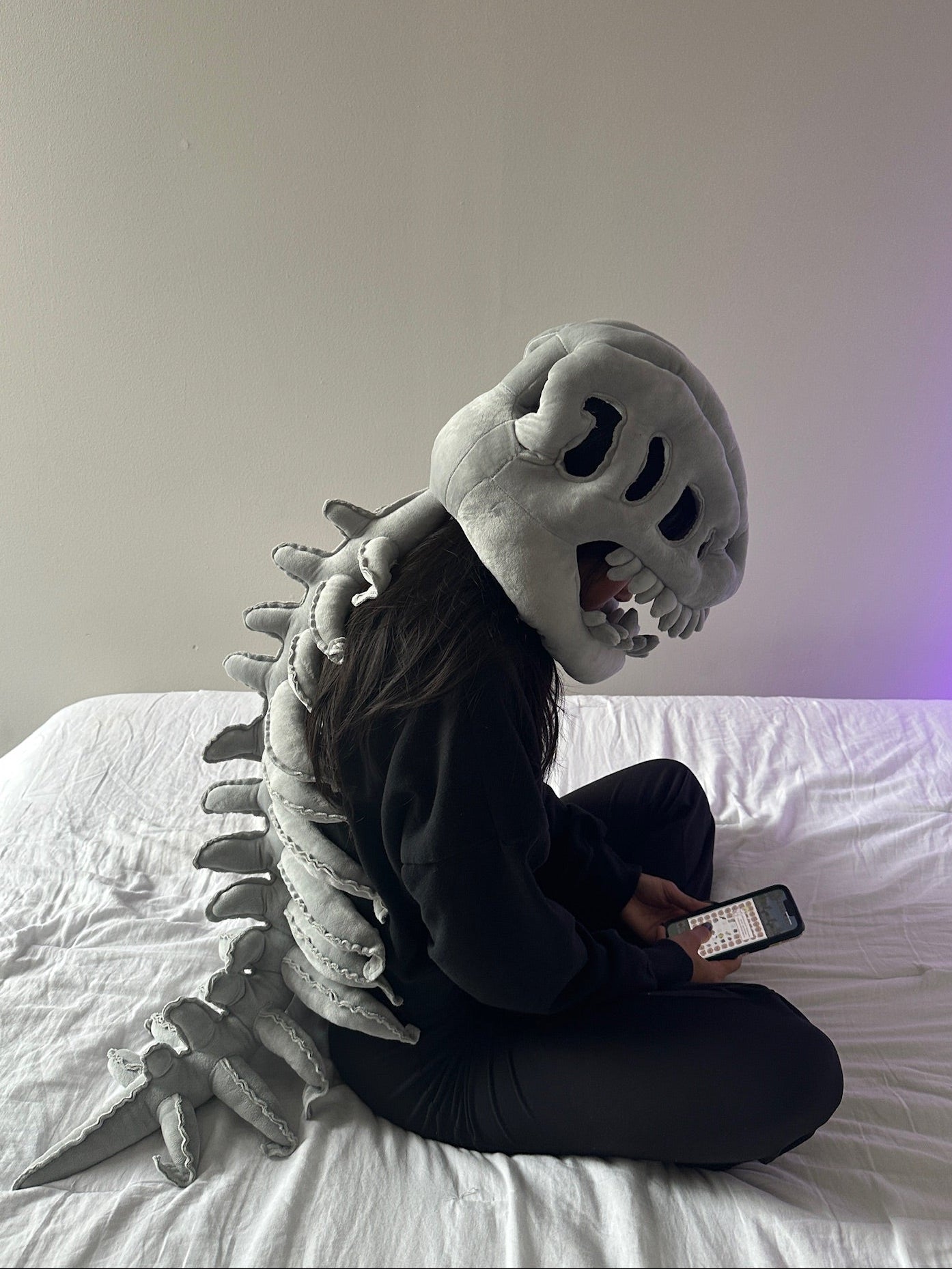 Person wearing a dinosaur Plush Huggie™ sitting on a bed using a phone.